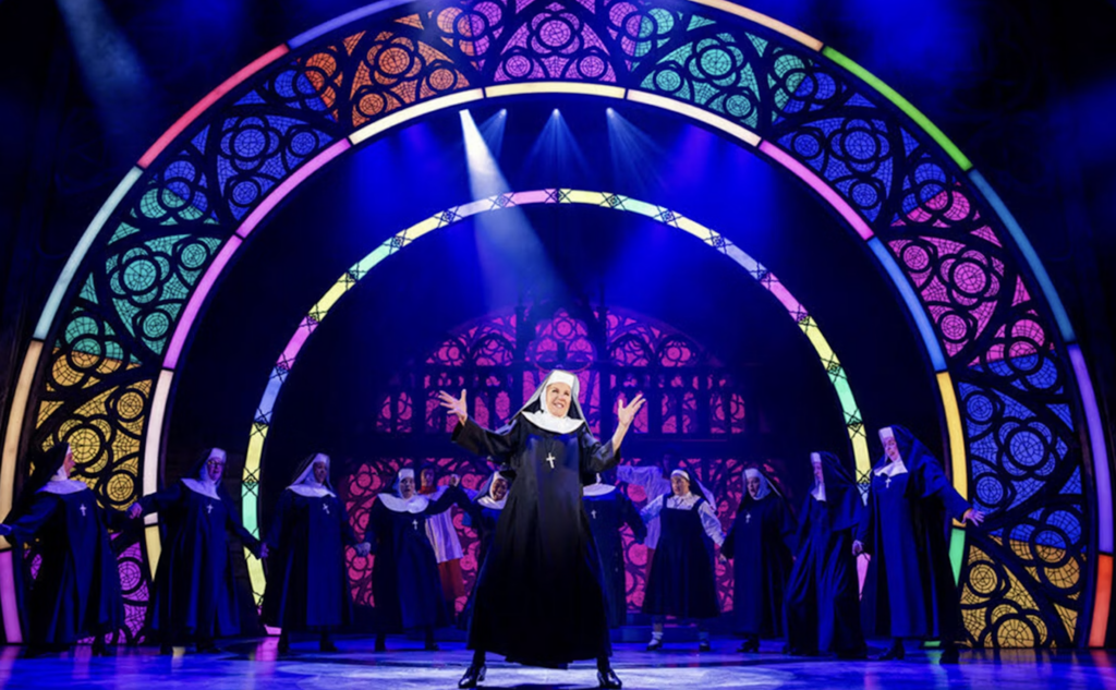 Rhonda Burchmore dances in a nun's habit in front of a chorus of nuns with a rainbow arch background.